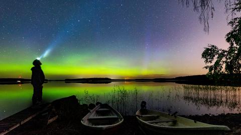 Yöllisessä venesatamassa seisoo henkilö, joka tähyää tähtitaivaalle otsalampullaan, kaupungin valot siintävät taivaanrannassa vihreänä ja violettina.